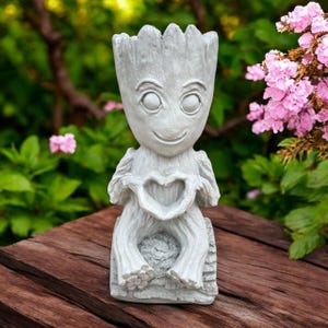 May include: A light gray, textured planter shaped like a humanoid tree character with large eyes and a smiling mouth. The figure is seated, with its hands positioned in front of its chest to form a heart shape. The top of the head is open.