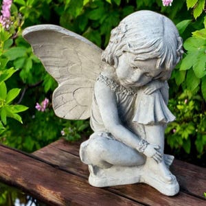 May include: A gray concrete fairy statue with large detailed wings, seated with its head bowed. The fairy is wearing a dress and bracelet. The statue is on a wooden surface, surrounded by green foliage and pink flowers.