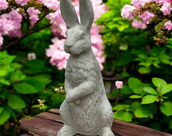 Può includere: Una statua di coniglio in cemento grigio con lunghe orecchie e una superficie strutturata. Il coniglio è in piedi con le zampe unite. La statua è posta su una superficie di legno, con piante fiorite rosa sullo sfondo.