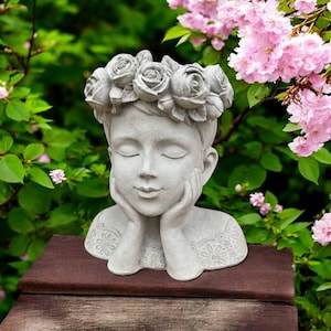 May include: A light gray stone-colored planter shaped like a human head with closed eyes and hands resting against the cheeks. The figure wears a crown of sculpted roses and a textured garment with a floral pattern across the chest and shoulders.