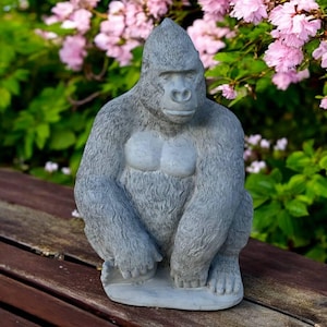 May include: A gray gorilla statue with a textured fur-like appearance, seated on a wooden surface. The gorilla has a light gray chest and is surrounded by green foliage and pink flowers, creating a naturalistic setting.