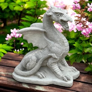 May include: A detailed, gray dragon statue with outstretched wings and an open mouth. The mythical creature is made of stone and sits on a stone base. The dragon is placed on a wooden surface, with a backdrop of green foliage and pink flowers.