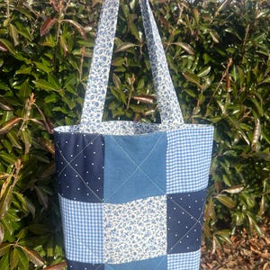 May include: A patchwork tote bag with a variety of blue fabrics. The bag features squares of denim, gingham, and floral patterns. The handles and interior lining have a blue floral design. The bag is held up by a person's hand.