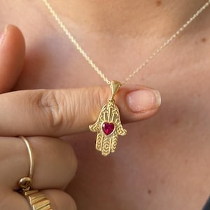 May include: A gold-toned Hamsa pendant necklace featuring an intricate engraved pattern and a small, central red heart-shaped stone. The pendant hangs from a delicate gold-toned chain, complemented by a wide, textured gold-toned band ring on a finger.