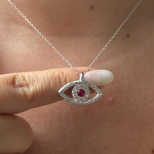 May include: A silver necklace featuring an eye-shaped pendant with a textured metallic finish. The pendant is encrusted with small clear stones surrounding a single, central round red stone, suspended from a delicate silver chain.