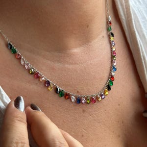 Peut inclure: Un collier en argent avec une chaîne fine ornée d'une série de pierres précieuses colorées en forme de goutte. Les pierres sont de différentes couleurs, dont le rouge, le vert, le bleu, le jaune et le rose. Le collier est porté autour du cou.