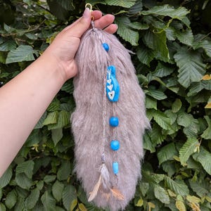 Può includere: Un portachiavi a coda in pelliccia sintetica marrone chiaro e soffice con una chiusura in metallo dorato. L'accessorio è decorato con un filo verticale di perline blu brillante, inclusa una perla più grande con un motivo a foglia bianca, e due piccole piume marroni.