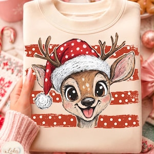 May include: A light beige crewneck sweatshirt featuring a graphic illustration of a reindeer wearing a red and white polka dot Santa hat. The reindeer has large dark eyes, a small nose, and a smiling mouth with its tongue visible.