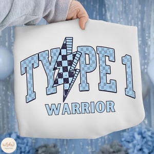 May include: White tote bag with the text "TYPE 1 WARRIOR" in light blue block letters. A blue and black checkered lightning bolt graphic is between "TYPE 1" and "1". The bag is held by a person.