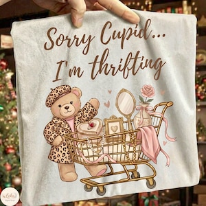 May include: A light gray pillow cover with the text "Sorry Cupid... I'm thrifting." A cartoon teddy bear in a leopard print coat and beret pushes a shopping cart filled with jewelry, a mirror, and a vase with a rose.