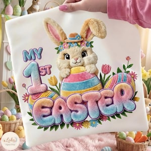 Könnte beinhalten: Weißes Ostermotiv mit einem Hasen, der einen Hut und einen Blumenkranz trägt und ein buntes Ei hält. Der Text lautet "MY 1ST EASTER" in Pastellfarben, umgeben von Blumen und Ostereiern. Ein festliches Design.
