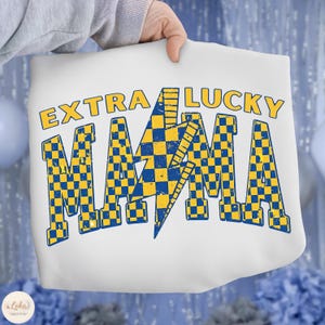 May include: White pillow with the text "EXTRA LUCKY MAMA" in yellow and blue. The word "MAMA" is in a checkered pattern with a blue lightning bolt in the center. The pillow is held by a person.
