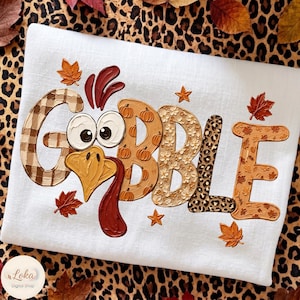 May include: A white t-shirt featuring the text GOBBLE in large letters. The G has a brown plaid pattern, the O is replaced by a turkey face, the B has a pumpkin print, the B has a floral pattern, the L has a leopard print, and the E has leaves.