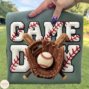 以下が含まれることがあります： 「GAME DAY」の文字が白で野球をテーマにしたデザインの緑色のスウェットシャツ。茶色と白のかぎ針編みの野球ボール、グローブ、バットが含まれています。スウェットシャツは手で持たれています。