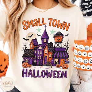 May include: A cream-colored t-shirt featuring a graphic of a spooky town with purple and orange buildings, a water tower, pumpkins, bats, and a full moon. The text reads "SMALL TOWN" in orange and "HALLOWEEN" in purple with two light green stars.