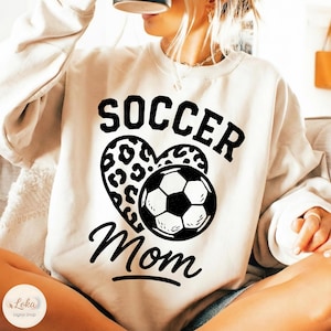 Puede incluir: Sudadera color crema con las palabras "SOCCER Mom" en negro. El diseño incluye un balón de fútbol y un corazón con estampado de leopardo. La sudadera es de estilo casual.