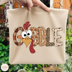 Könnte beinhalten: Eine beige Tragetasche mit dem Wort "GOBBLE" in einem Patchwork-Design und einem Truthahn-Gesicht. Die Buchstaben sind mit Karomuster, Kürbissen, Leopardenmuster und Herbstblättern verziert. Der Truthahn hat große Augen und einen roten Wamm.