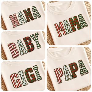 May include: Cream-coloured sweatshirts featuring embroidered text in festive patterns. The words "NANA", "MAMA", "BABY", "GIGI", and "PAPA" are displayed in red and green stripes, plaid, and Christmas tree designs, ideal for holiday gifting.