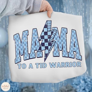 Puede incluir: Sudadera blanca con el texto "MAMA TO A T1D WARRIOR" en azul claro, fuente desgastada. La palabra "MAMA" está en un patrón de cuadros con un rayo azul y negro en el centro. Un gran regalo para una madre.