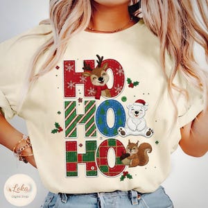 Könnte beinhalten: Cremefarbenes Sweatshirt mit festlichem "HO HO HO" Design. Die Buchstaben sind in rot, grün und blau karierten Mustern gehalten, mit Illustrationen von Rentier, Eisbär und Eichhörnchen. Enthält Schneeflocken und Stechpalmen.