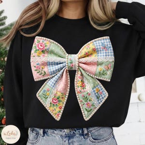 May include: Black sweatshirt featuring a large, colorful bow design. The bow is made of patchwork fabric with floral, polka dot, and gingham patterns. It is trimmed with lace and faux pearls, and has embroidered butterflies.