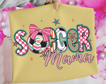 Puede incluir: Un cojín amarillo claro con las palabras "SOCCER Mama" en un diseño colorido. La palabra "SOCCER" tiene un balón de fútbol en el centro con un lazo rosa. El cojín está sostenido por una persona.