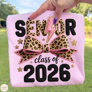 Puede incluir: Una sudadera rosa claro con la palabra "SENIOR" en letras negras y con estampado de leopardo, un rayo rosa y un lazo con estampado de leopardo. El texto "class of 2026" está debajo del lazo, con estrellas.