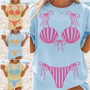 May include: A light blue t-shirt featuring a pink seashell bikini design. The bikini top and bottom are outlined with bows. The design is a fun, summery graphic, perfect for beach or pool wear.