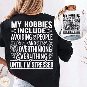 Puede incluir: Sudadera negra con texto blanco: "MY HOBBIES INCLUDE AVOIDING PEOPLE AND OVERTHINKING EVERYTHING UNTIL I'M STRESSED." Una imagen más pequeña muestra una sudadera blanca con el mismo texto. Es una prenda casual y cómoda.