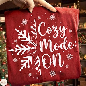 May include: A red pillow with white snowflakes and the words "Cozy Mode: ON" in a script font. The pillow is held up by a hand, suggesting it's a decorative item for the home, perfect for the holiday season.