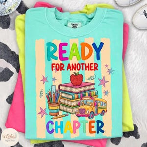 Może przedstawiać: Jasnoturkusowy t-shirt z nadrukiem graficznym z napisem READY FOR ANOTHER CHAPTER. Wzór przedstawia stos książek z czerwonym jabłkiem na szczycie, kubek z kredkami oraz żółty autobus szkolny, a wszystko to otoczone jest gwiazdkami.