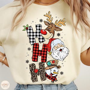 Könnte beinhalten: Cremefarbenes T-Shirt mit dem Schriftzug "HO HO HO" in einem festlichen Design. Die Buchstaben sind mit einem Rentier, dem Weihnachtsmann und einem Schneemann verziert. Die Buchstaben "H" haben Karomuster und Leopardenmuster. Schneeflocken sind um das Design herum verstreut.