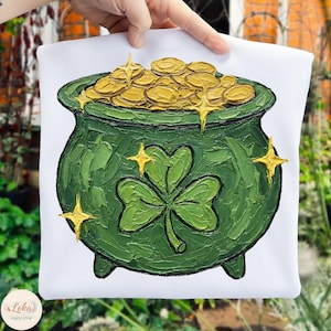 Puede incluir: Camiseta blanca con un diseño de olla de oro verde, un trébol y monedas de oro. El diseño tiene un aspecto texturizado y pintado con acentos de estrellas doradas. Un diseño festivo para el Día de San Patricio.