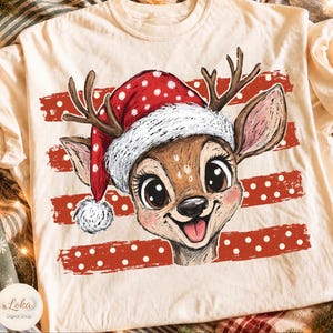 May include: A cream-colored t-shirt featuring a graphic illustration of a deer wearing a red and white polka-dot Santa hat. The deer has large eyes, antlers, and a smiling expression. Behind the deer are three horizontal red, distressed stripes.