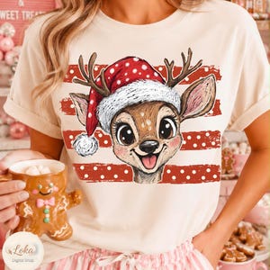 May include: A cream-colored t-shirt featuring a graphic of a brown deer wearing a red and white polka-dot Santa hat. The deer is centered over horizontal red stripes with white polka dots. The design includes large eyes and a smiling mouth.