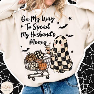 May include: A cream-colored sweatshirt featuring a graphic of a ghost in a checkered sheet pushing a shopping cart filled with pumpkins. The text reads, On My Way To Spend My Husband's Money, accompanied by small black bat and star illustrations.