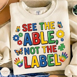May include: Cream-colored sweatshirt with the text "SEE THE ABLE, NOT THE LABEL" in colorful block letters. The design includes butterflies, gears, puzzle pieces, and a rainbow handprint. A casual, comfortable top.