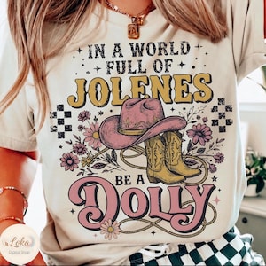 Puede incluir: Una camiseta color crema con un estampado gráfico que incluye el texto IN A WORLD FULL OF JOLENES BE A DOLLY. El diseño presenta un sombrero de vaquero rosa, botas amarillas, un lazo y flores moradas, acentuado con cuadrados negros a cuadros.