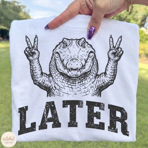 May include: White t-shirt featuring a black and white illustration of an alligator making a peace sign with its hands. The word "LATER" is printed in large, distressed black letters below the alligator image. The shirt is held up against a green background.
