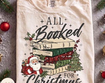 May include: A cream-colored t-shirt featuring a graphic of a stack of books, a Santa Claus figure, and a Christmas tree. The text reads ALL Booked FOR Christmas. The design includes holly leaves, berries, and small star accents in black and red.