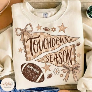 Pode incluir: Uma camisola de gola redonda cor de creme com uma impressão gráfica castanha. O design inclui duas bandeirolas axadrezadas com o texto "TOUCHDOWN SEASON", dois laços, várias estrelas, pequenas flores e três bolas de futebol americano.