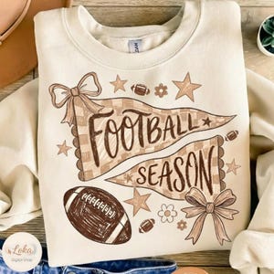 May include: A cream-colored crewneck sweatshirt featuring a brown graphic print of two pennants with the text "FOOTBALL SEASON". The design includes illustrations of a football, stars, flowers, and two bows in a brown and tan color palette.