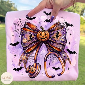 May include: A light purple t-shirt featuring a graphic print of a large bow with a central jack-o'-lantern, integrated with a stethoscope, spiderwebs, flying bats, silhouettes of witches on broomsticks, and small potion bottles in orange and purple.