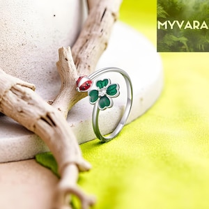 Puede incluir: Un anillo plateado con un diseño de trébol de cuatro hojas verde y blanco y un detalle de mariquita roja y blanca. El anillo está sobre un fondo amarillo-verdoso brillante, con una rama de madera en primer plano. El nombre de la marca "MYVARA" es visible.