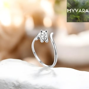 May include: A silver ring with a cat design. The cat is lying on the ring's band, with its tail curved upwards. The ring is set against a soft, blurred background. The ring is a piece of jewellery.