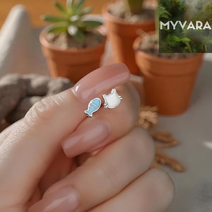 Puede incluir: Un par de pendientes de plata. Un pendiente es un gato blanco con detalles plateados, y el otro es un pez azul y plateado. Los pendientes son pequeños y delicados.