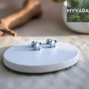 May include: A pair of silver stud earrings. Each earring has a detailed design on the front and a smooth, round backing. The earrings are presented on a light blue, circular surface. The earrings are likely made of metal.