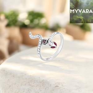 May include: A silver snake ring with a curved design. The ring features a snake head with a red gemstone and a body adorned with small, clear gemstones. The ring is set on a white surface. The brand name "MYVARA" is visible in the background.