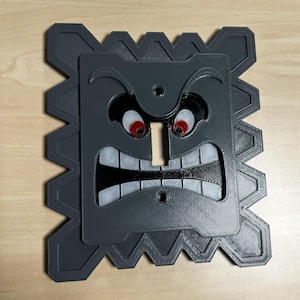 May include: A gray, 3D-printed light switch cover featuring a cartoonish face with red eyes, a wide open mouth, and jagged edges. The design is reminiscent of a character from a popular video game.