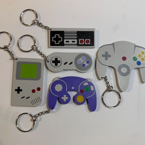 May include: A collection of five video game themed keychains featuring a gray handheld console, a rectangular controller, a rounded controller, a purple ergonomic controller, and a gray three-pronged controller, each attached to a metal split ring.